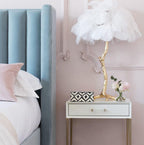 White-feathered Clérachic table lamp on a brass branch stem, on a white bedside table with a patterned box and pink flowers.