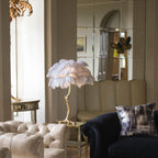 Clérachic feather lamp with pale white-lilac plumes on a gold side table between a cream button-back sofa and a dark armchair.
