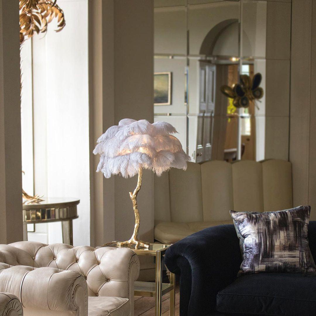 Clérachic feather lamp with pale white-lilac plumes on a gold side table between a cream button-back sofa and a dark armchair.