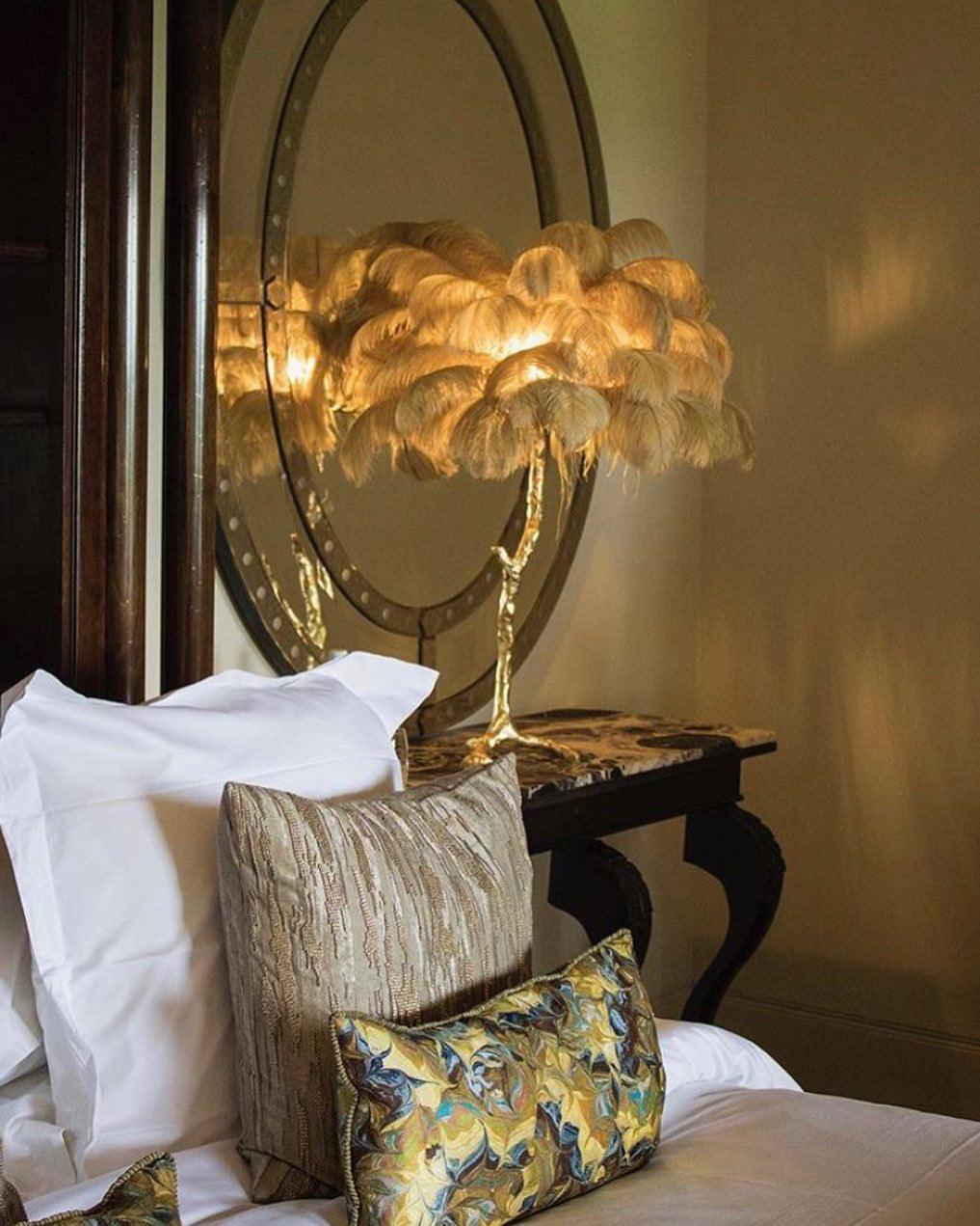 Lit Clérachic feather lamp with golden plumes beside white and patterned pillows, with an oval mirror behind in warm light.