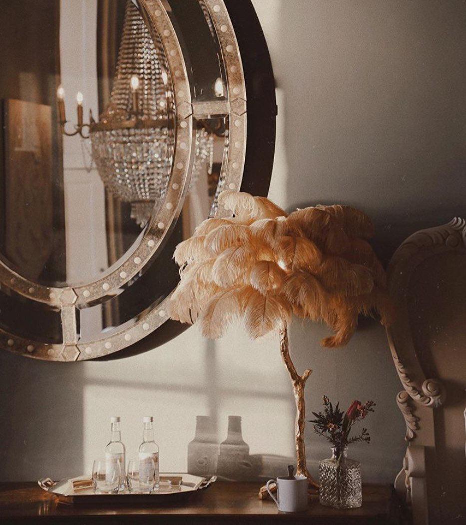 Clérachic feather lamp with golden-tan plumes on a console, with a round studded mirror, a chandelier and a tray of glasses behind.