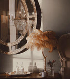 Clérachic feather lamp with golden-tan plumes on a console, with a round studded mirror, a chandelier and a tray of glasses behind.