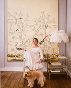 A white-feathered Clérachic table lamp on a gold side table beside a seated woman and a poodle, in front of a bird-and-blossom mural.