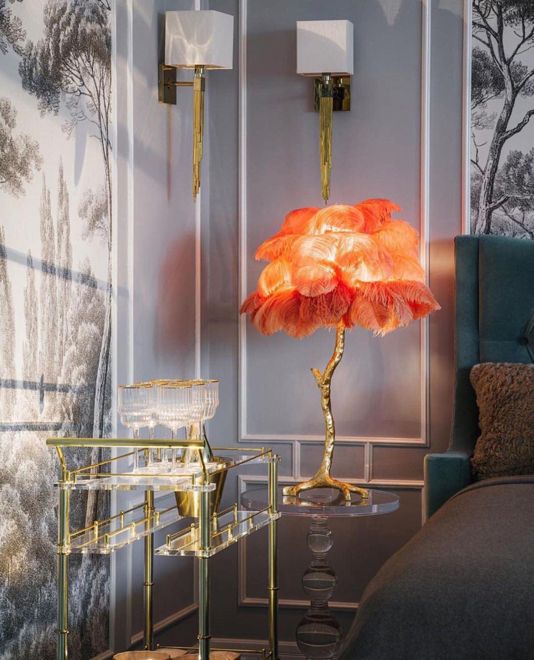 Glowing orange Clérachic feather lamp on a brass stem beside a drinks trolley, with two wall lights and tree-print wallpaper.