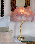 Close view of a Clérachic feather lamp with pink plumes lit up, on a glass table by a carved bedpost and a gilt mirror.