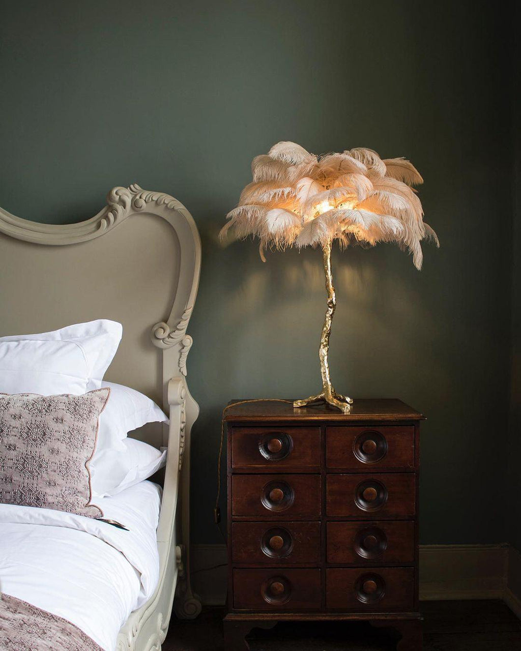 A Clérachic table lamp with peach feathers lit on a chest of drawers, beside a cream bed with white pillows against a dark green wall.