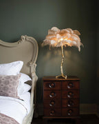 A Clérachic table lamp with peach feathers lit on a chest of drawers, beside a cream bed with white pillows against a dark green wall.