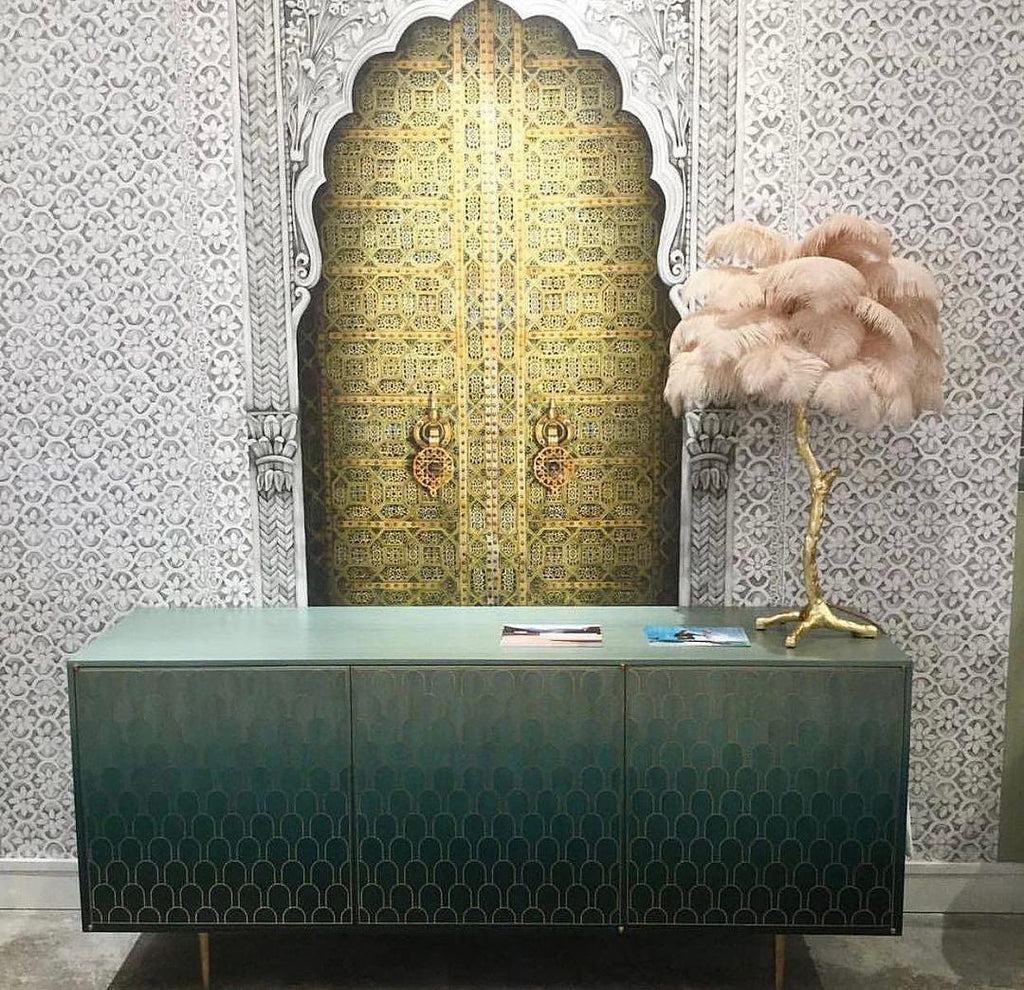 Clérachic feather lamp with pale peach plumes on a green sideboard, in front of a gold arched door and patterned wallpaper.
