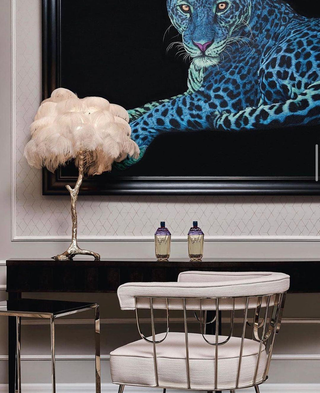 Cream-feathered Clérachic table lamp on a dark console in front of a painting of a blue leopard, with a metal-framed chair below.