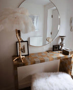 A white Clérachic feather lamp on a gold branch stem on a mosaic-tiled dressing table, with a round mirror and a fluffy stool.