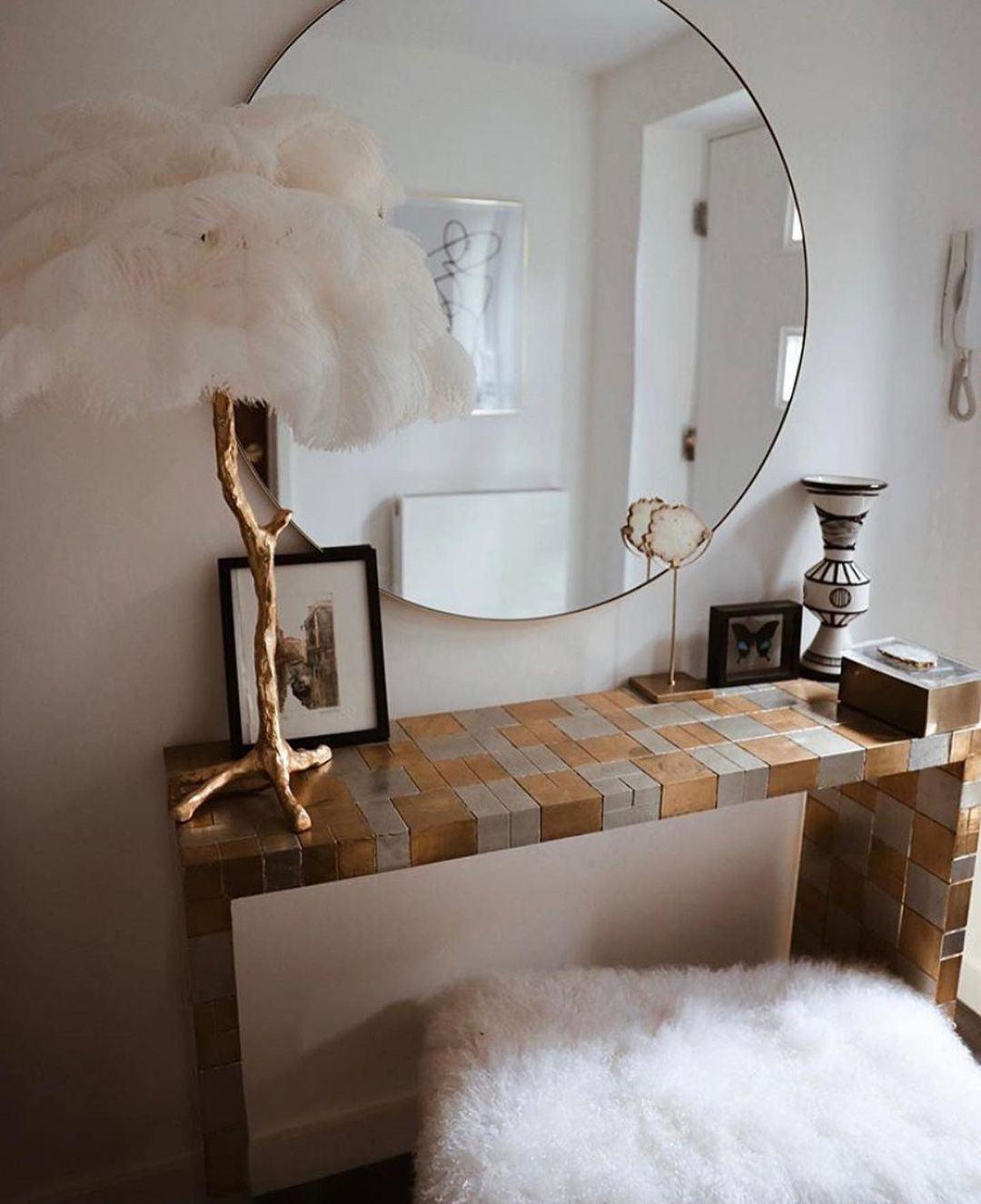 A white Clérachic feather lamp on a gold branch stem on a mosaic-tiled dressing table, with a round mirror and a fluffy stool.