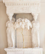 Clérachic table lamp with white feathers on a gold stem on a stone mantel between two carved caryatid figures.