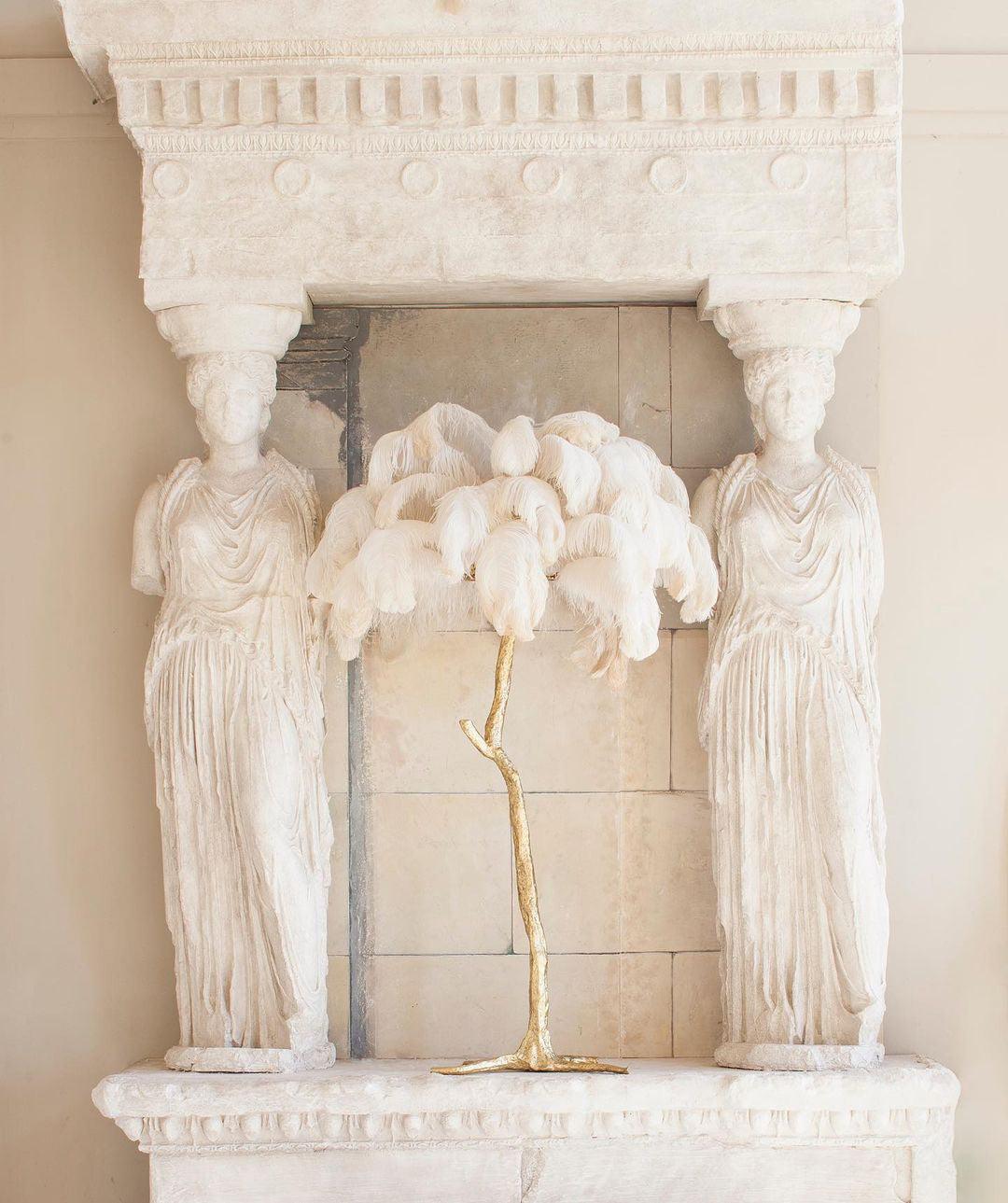Clérachic table lamp with white feathers on a gold stem on a stone mantel between two carved caryatid figures.