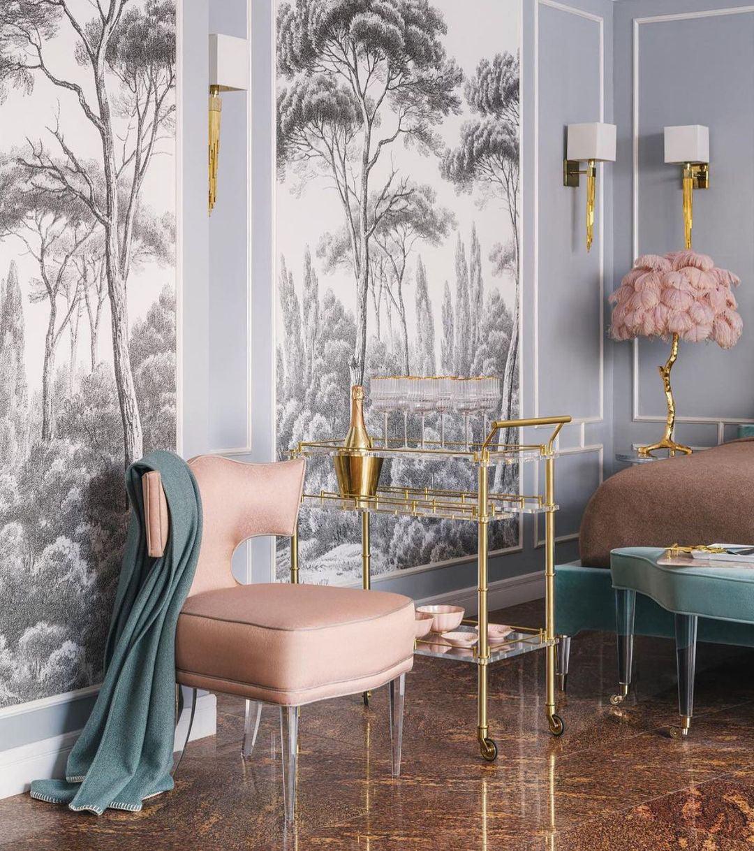 A Clérachic feather lamp with pink plumes in the distance of a room with tree-print wallpaper, a pink chair and a brass drinks trolley.