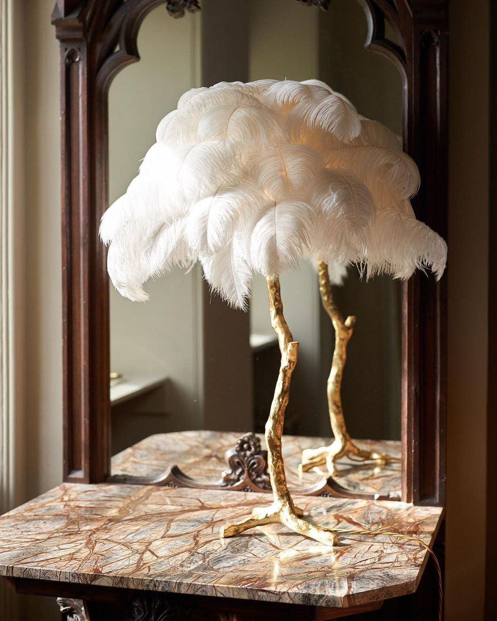 Clérachic table lamp with large white feathers on a gold stem, on a marble-topped table in front of a dark wood mirror.