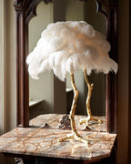 Clérachic table lamp with large white feathers on a gold stem, on a marble-topped table in front of a dark wood mirror.