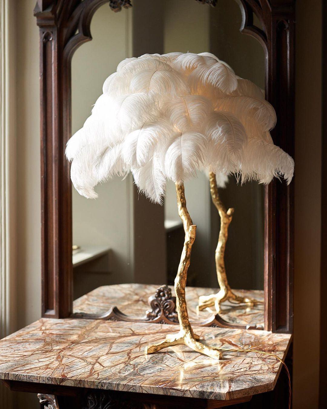 Clérachic table lamp with large white feathers on a gold stem, on a marble-topped table in front of a dark wood mirror.