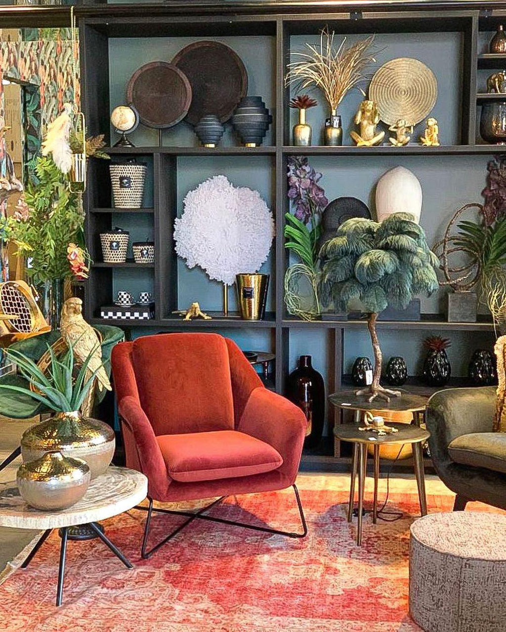 A Clérachic table lamp with grey-green feathers among shelves of ornaments, with a red armchair and a patterned rug in front.