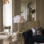 Clérachic table lamp with pale lilac-white feathers on a gold side table beside cream button-back sofas and a navy armchair.