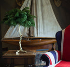 Close view of the Clérachic table lamp with dark green feathers on a wooden table, with a model boat and a Union Jack chair behind.