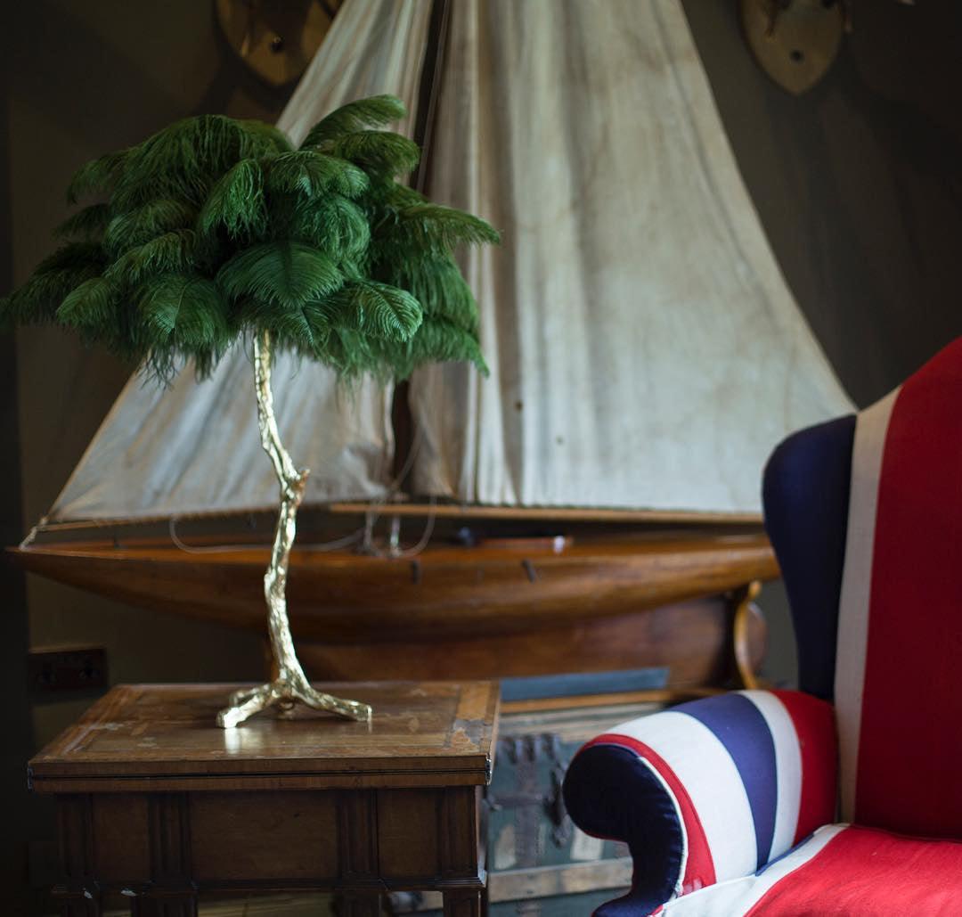 Close view of the Clérachic table lamp with dark green feathers on a wooden table, with a model boat and a Union Jack chair behind.