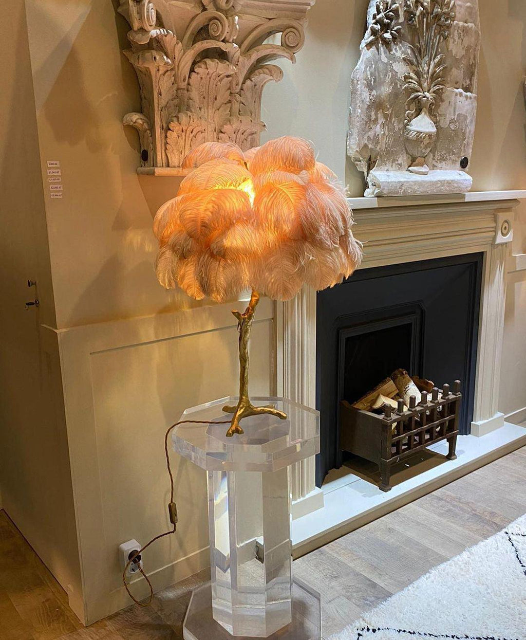 Clérachic table lamp with peach plumes glowing on a clear acrylic stand, in front of a marble fireplace with a stone capital above.