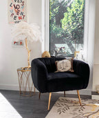 A white-feathered Clérachic table lamp on a gold side table, beside a black velvet armchair and a window with a view of trees.