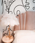 Clérachic feather lamp with white plumes on a gold branch stem, on a round bedside table in front of a blush headboard and a line-art wall.