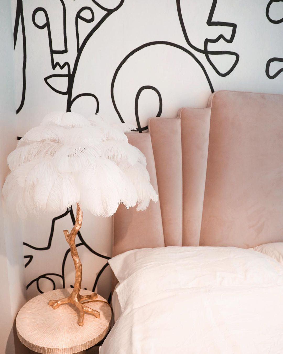 Clérachic feather lamp with white plumes on a gold branch stem, on a round bedside table in front of a blush headboard and a line-art wall.