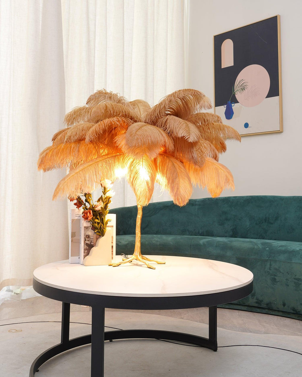 Lit Clérachic feather lamp with camel-gold plumes on a round table with a vase of flowers, in front of a green velvet sofa.