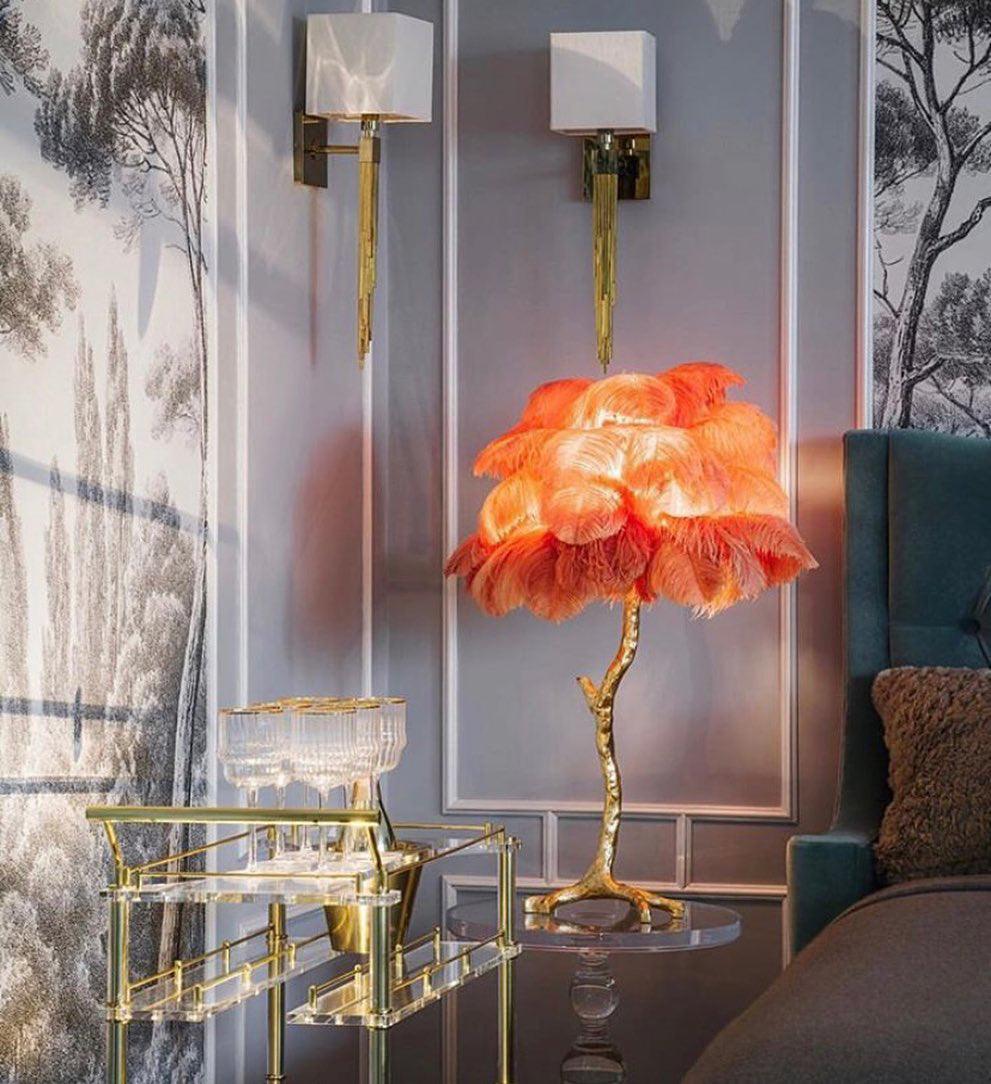 Glowing orange Clérachic feather lamp on a gold stem, beside a brass drinks trolley, a teal headboard and two wall lights.