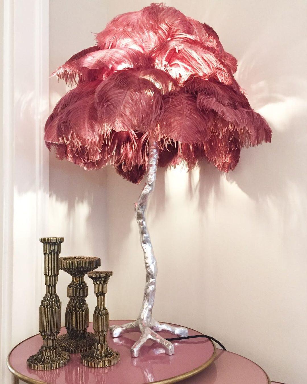 Clérachic table lamp with deep raspberry feathers on a silver branch stem, on a pink table beside three brass candlesticks.