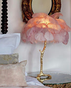 Clérachic feather lamp with soft pink plumes lit up, on a glass table beside cushions, a carved bedpost and a gilt mirror.