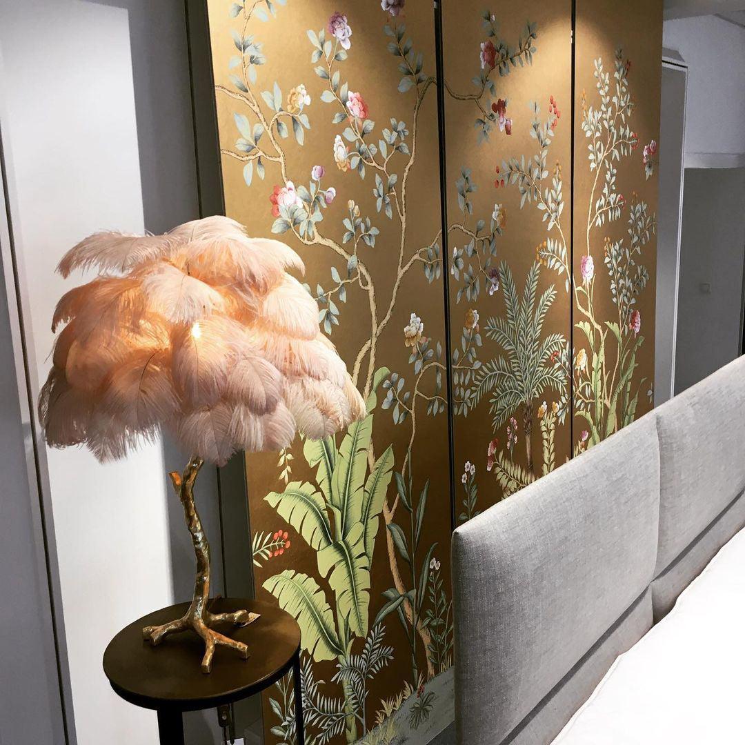 Clérachic feather lamp with peach plumes on a small black table, in front of a gold mural of leaves and blossom and a grey headboard.