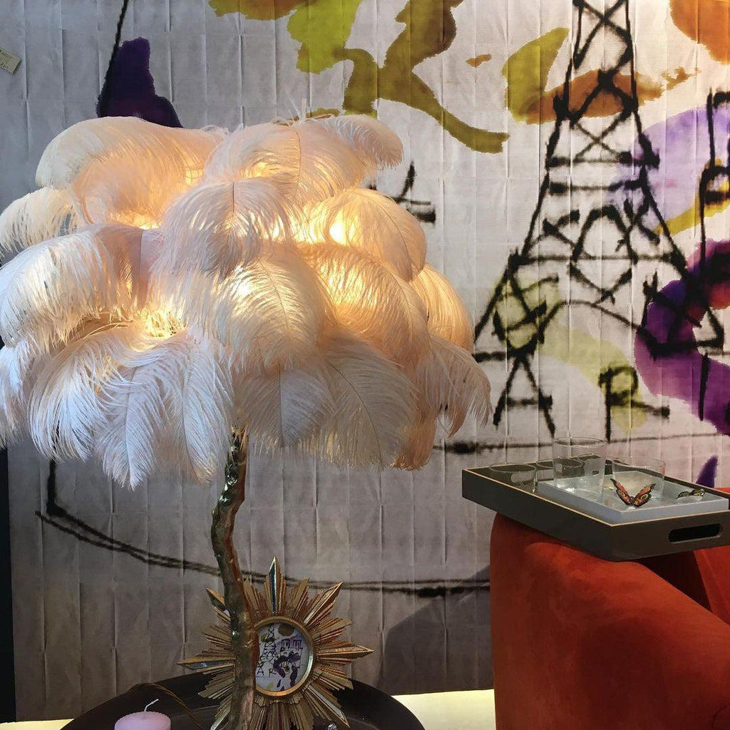 Close view of a Clérachic feather lamp with soft white-pink plumes lit up, beside a sunburst mirror and a rust-coloured chair.