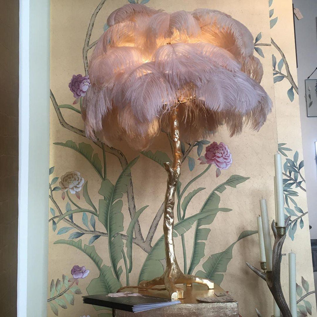 Clérachic feather lamp with dusky pink plumes lit up, in front of a gold wall mural of peonies and banana leaves.