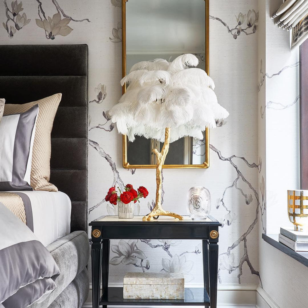 A white Clérachic feather lamp on a gold branch stem on a black side table, in front of a gilt mirror and blossom wallpaper.