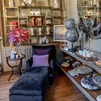 Clérachic table lamp with plum-purple feathers on a shelf in a book-lined room, beside a dark velvet chaise and a pair of globes.