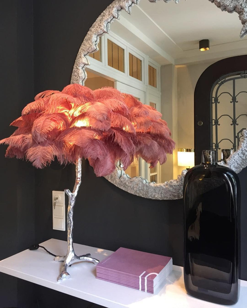 Lit Clérachic feather lamp with coral-pink plumes on a silver stem, on a white shelf below a round textured mirror and a black glass vase.