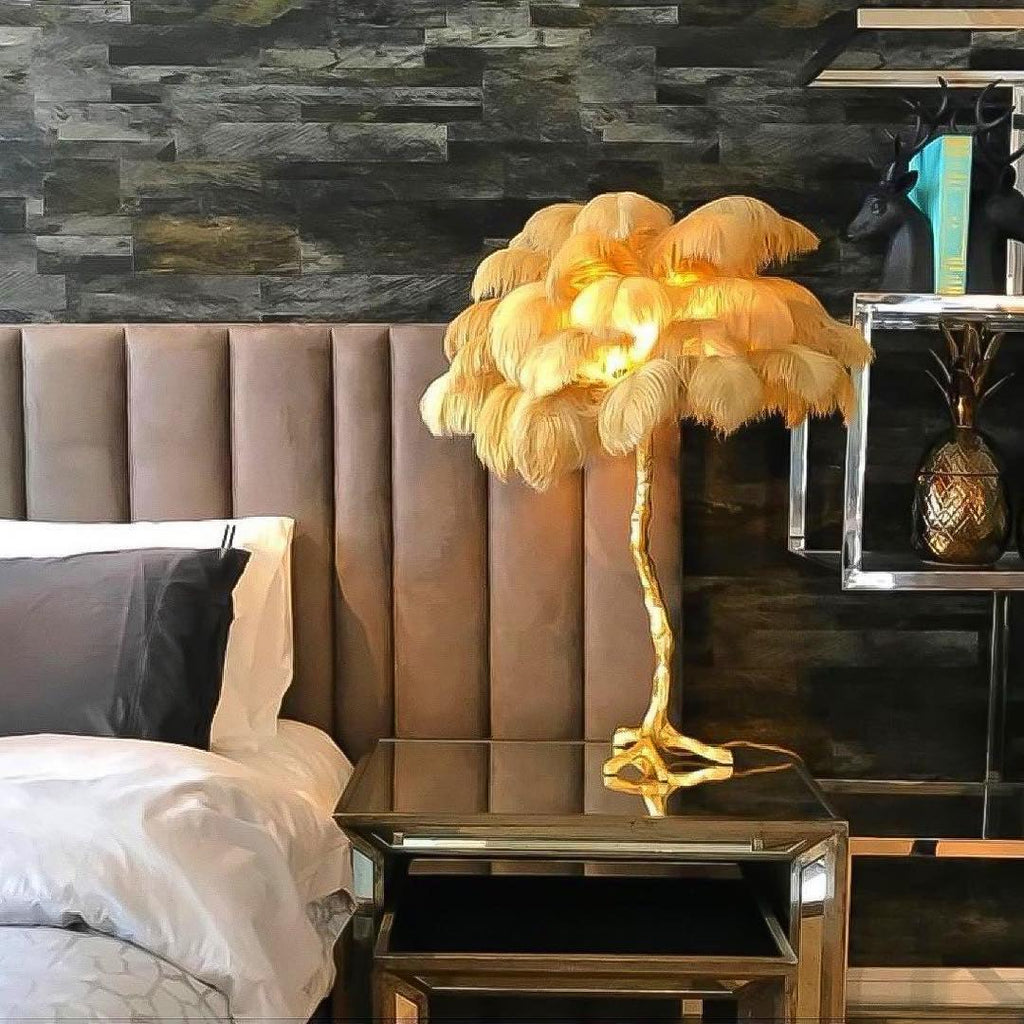 Lit Clérachic feather lamp with golden plumes on a mirrored bedside table, beside a velvet panelled headboard and a brass pineapple.