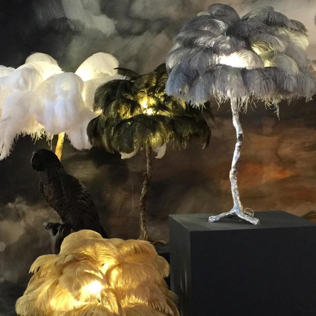 Clérachic feather lamp with grey plumes on a silver stem on a black plinth, with white and golden feather lamps glowing behind and beside it.