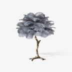 Clérachic table lamp with grey layered ostrich feathers on a gold branch stem, seen on a plain light backdrop with a soft shadow.