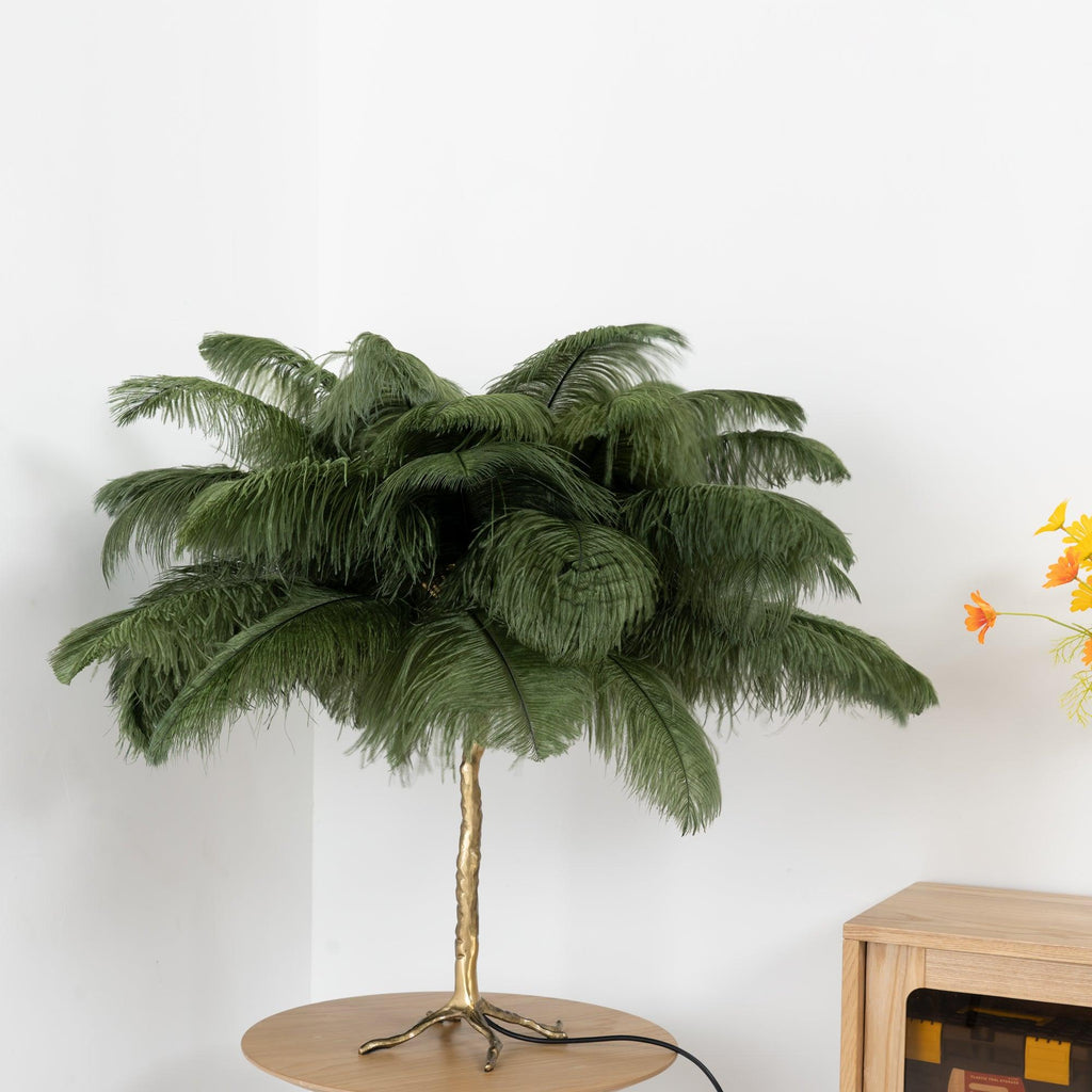 Clérachic table lamp with an olive-green feather shade on a gold branch stem, on a round wooden table beside a small wooden cabinet.