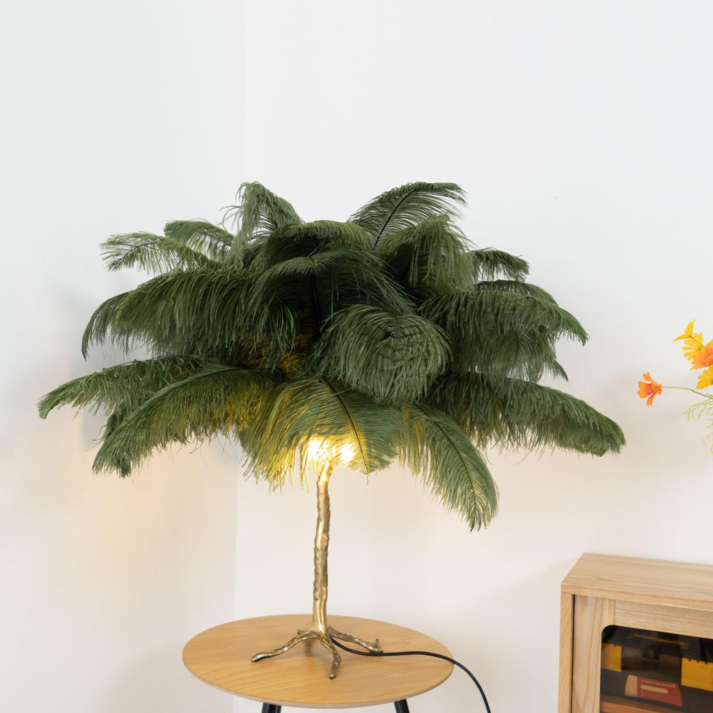 Lit Clérachic feather lamp with olive-green plumes and a warm glow at the base, on a round wooden table in a pale room.