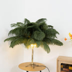 Lit Clérachic feather lamp with olive-green plumes and a warm glow at the base, on a round wooden table in a pale room.