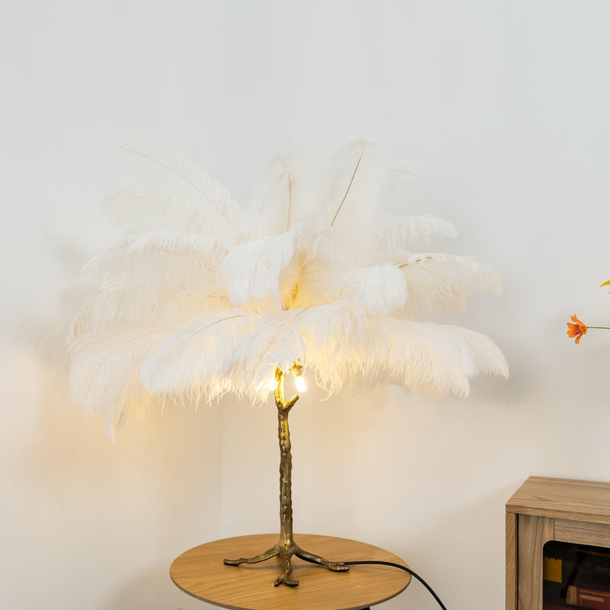 Clérachic feather lamp with white plumes lit from below on a round wooden table, in front of a pale wall and a wooden cabinet.