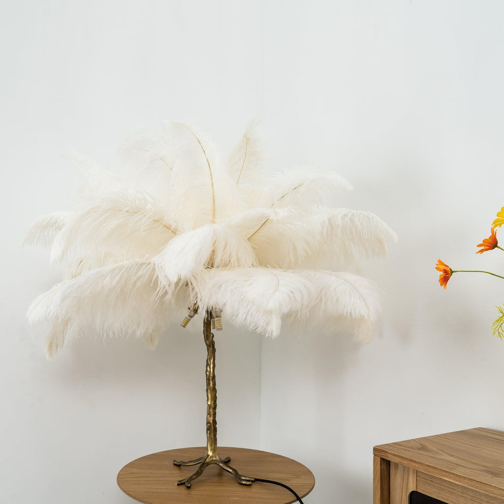 Clérachic table lamp with unlit cream-white feathers on a gold branch stem, on a round wooden table by a pale wall.