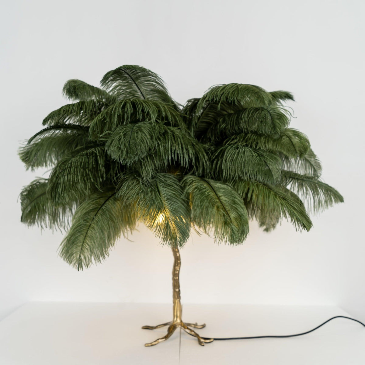 Front view of a Clérachic table lamp with dark green feathers and a warm glow at the stem, on a white surface with a black cord.
