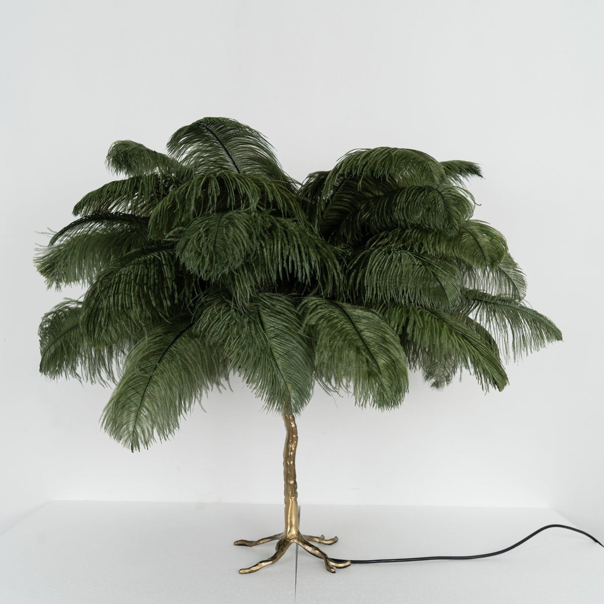 Clérachic table lamp with unlit dark green feathers on a gold branch stem, shown on a white shelf with its black cord trailing right.
