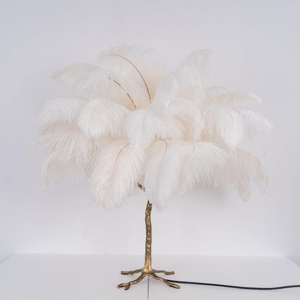Front view of a Clérachic table lamp with unlit white ostrich feathers on a gold branch stem, with its black cord on a white surface.